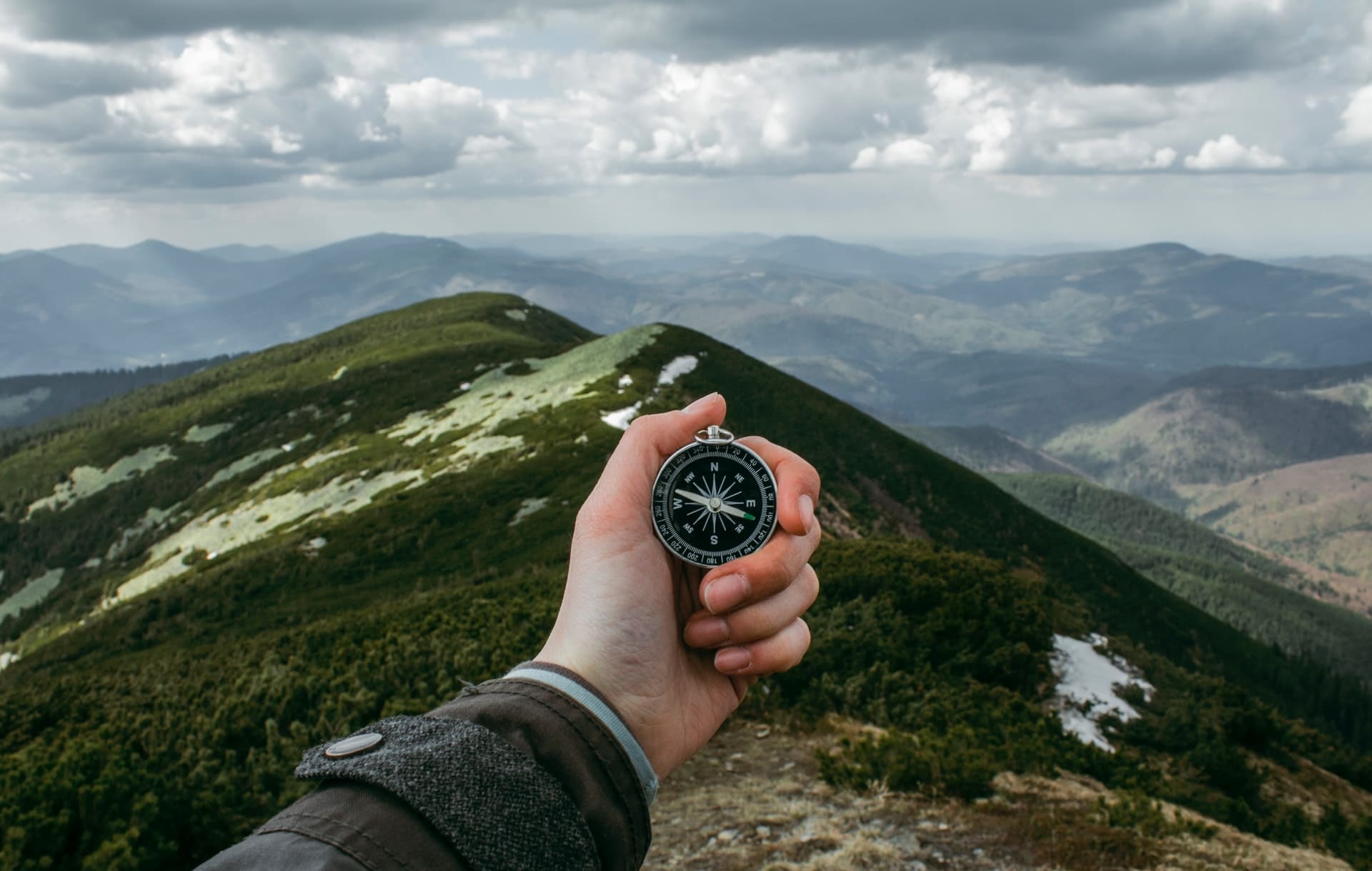 A compass held out in front of the camera with mountains and cloudy skies in the background.